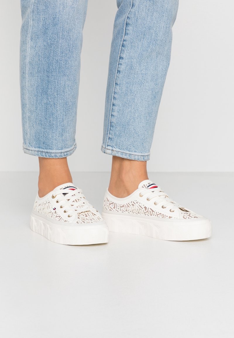 Persona che indossa sneakers platform bianche con motivo di pizzo e occhielli dorati, abbinate a jeans cropped azzurri, su uno sfondo semplice.