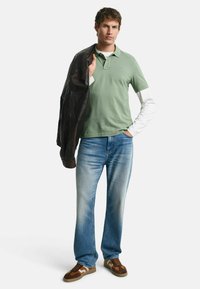 Camisa polo verde sobre una camisa de manga larga blanca, combinada con jeans azules y zapatillas deportivas marrones. Los accesorios incluyen una chaqueta de cuero negra.