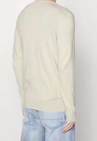 Jasnobeżowy sweter z okrągłym dekoltem i długimi rękawami, z prążkowanymi mankietami i dołem. Noszony z jasnoniebieskimi, luźno dopasowanymi dżinsami.