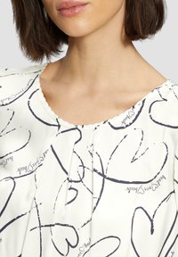 Blouse blanche avec des motifs de cœurs navy. Dispose d'un col rond avec une bordure ondulée et d'un tissu doux et léger. Les plis ajoutent de la texture.
