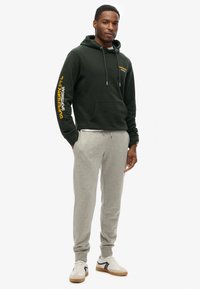 Felpa verde con logo sulla manica, pantaloni grigi e scarpe bianche con dettagli neri. Include cappuccio con cordino e tasca frontale.