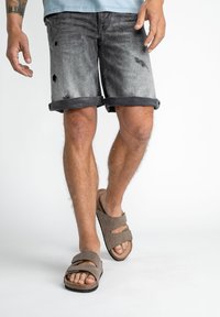 Grå denimshorts med slitna detaljer, vikta fållar, kombinerat med bruna sandaler med två remmar och en texturerad sula.