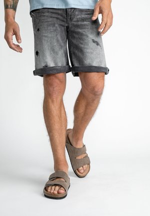 Graue Denim-Shorts mit distressed Details, umgeschlagenen Säumen, kombiniert mit braunen Sandalen, die zwei Riemen und eine strukturierte Sohle haben.