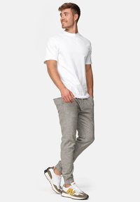 Camiseta blanca de manga corta, pantalones gris con textura y ajustados, y zapatillas blancas con detalles en gris y amarillo, de pie contra un fondo liso.
