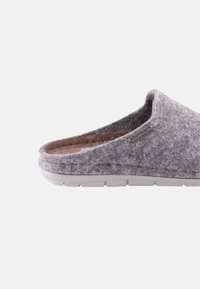 Grijze slipper van stof met een open achterkant, zachte textuur en een witte rubberen zool. Heeft een klein merklabel aan de zijkant.