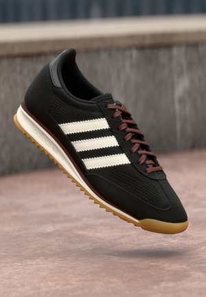 Czarny sneaker Adidas z białymi paskami, brązowymi sznurówkami i gumową podeszwą, pokazany w powietrzu na rozmytym tle kamienia i betonu.