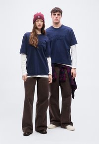 Δύο μοντέλα στέκονται φορώντας μπλουζάκια Tommy Jeans σε ναυτικό μπλε, πάνω από λευκά μακρυμάνικα, με καφέ παντελόνια φαρδιάς γραμμής και casual παπούτσια.