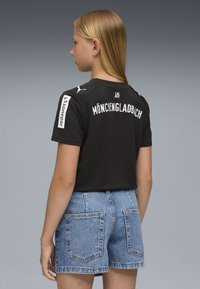 Giovane con capelli biondi che indossa una maglia sportiva nera del Mönchengladbach e pantaloni shorts in denim blu, in piedi contro uno sfondo grigio.