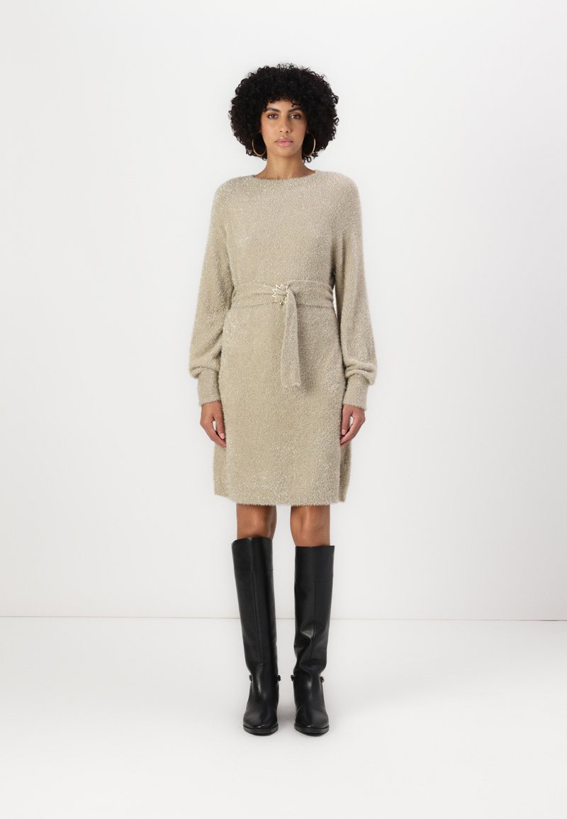 Beiges Strickkleid mit langen Ärmeln, Taillengürtel und glänzender Textur; kombiniert mit knielangen schwarzen Stiefeln. Schlichter runder Ausschnitt und minimale Details.