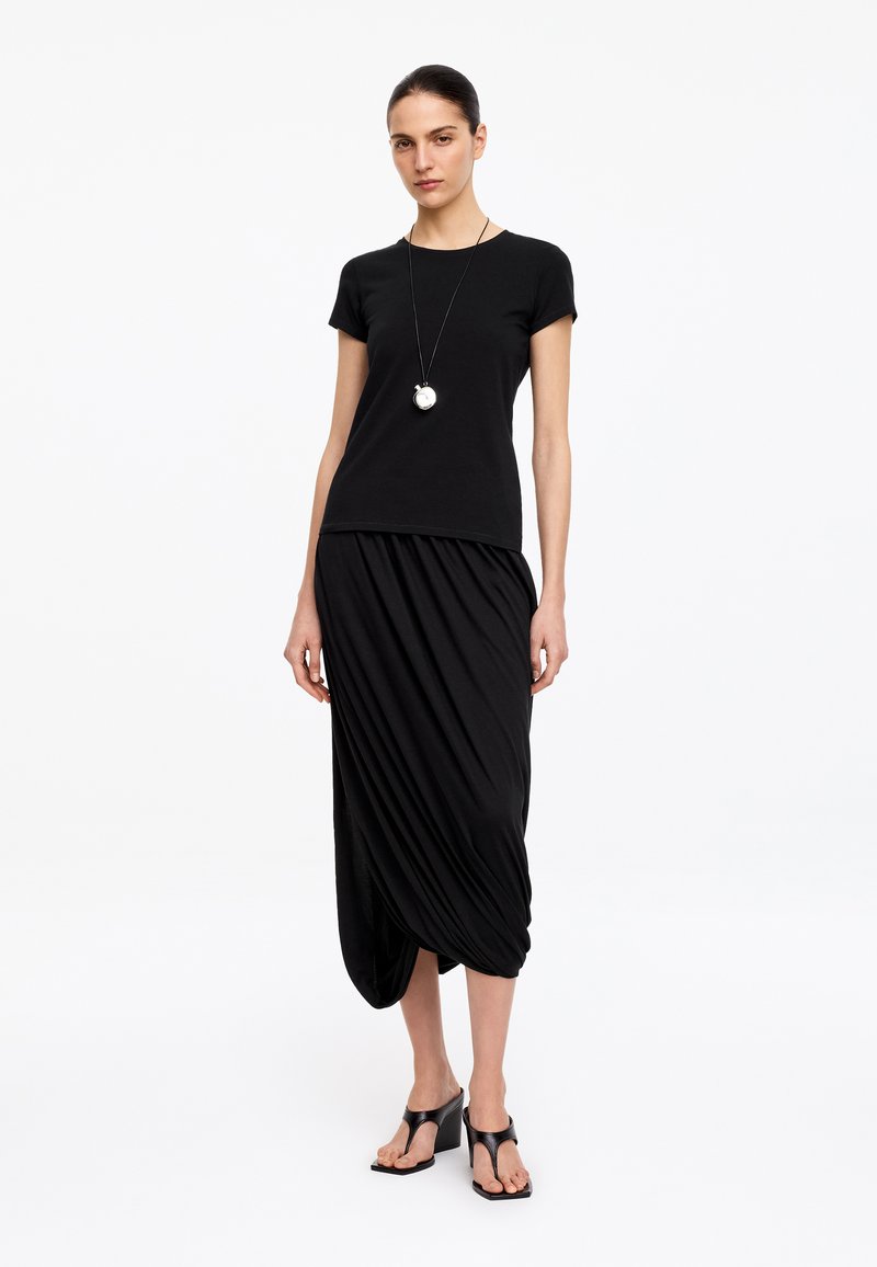 Femme portant un haut noir à manches courtes, une jupe midi noire drapée, des sandales noires à talons et un long collier avec un pendentif rond.
