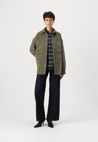 Casaco oversized verde em algodão com dois grandes bolsos frontais, sobre uma camisa xadrez preta e branca, combinado com jeans largos escuros e saltos pretos.