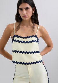Top corto en amarillo pálido con ribete ondulado en azul marino en el escote y el dobladillo. Presenta tirantes finos y un diseño de tela suelta y texturizada.