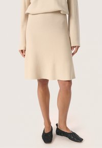 Beige strikket nederdel med flared kant, parret med sorte flade sko. Glat tekstur og enkelt design med elastisk talje.