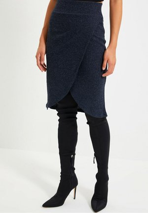 Vrouw draagt een donker geribbelde wikkelrok over zwarte kniehoge laarzen met puntige neus en hak, staand tegen een effen achtergrond.