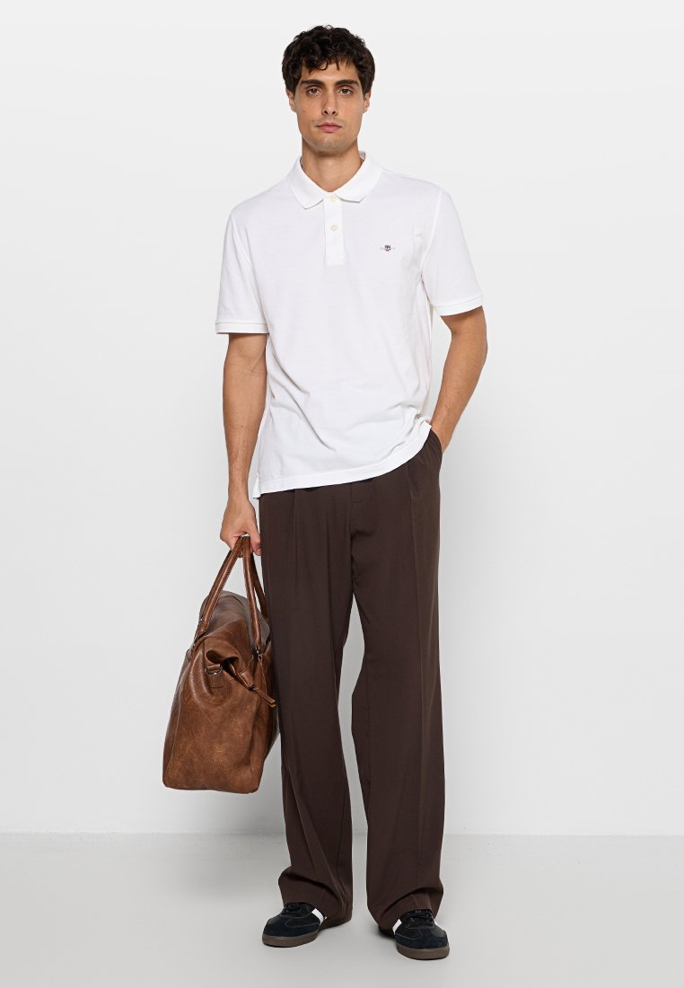 Weißes Polohemd mit kurzem Kragen, braune weit geschnittene Hose und braune Lederhandtasche. Schwarze Schuhe vervollständigen das Outfit.