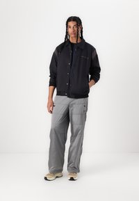 Chaqueta ligera negra con botones y bolsillos laterales, combinada con pantalones cargo grises que tienen bolsillos cargo y sutiles detalles de costura.