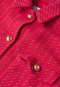 Czerwona kurtka z tweedu o teksturowanym wzorze, z złotymi guzikami i kołnierzem. Posiada kieszeń na piersi z klapą, pokazującą zbliżenie na splot materiału.