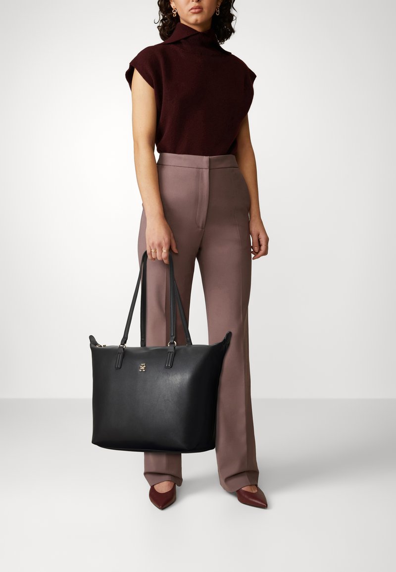 Borsa tote nera in pelle con due manici, con un emblema dorato sul davanti. Texture liscia, design spazioso e forma strutturata.