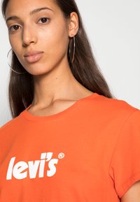 Camiseta de algodón naranja con un logo blanco de "levi's", cuello redondo y mangas cortas. Modelo lleva pendientes de aro plateados grandes.