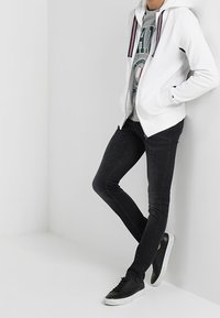 Sudadera blanca con cremallera y acentos en rayas rojas y azules sobre una sudadera gráfica gris, combinada con jeans negros de corte ajustado y zapatillas negras.