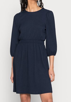 Kobieta ubrana w granatową sukienkę do kolan z rękawami 3/4 z bufkami i elastyczną talią, stojąca na tle jednolitego jasnego tła.