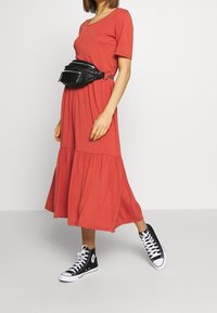 Ein rostfarbendes, knielanges Kleid mit kurzen Ärmeln und gestuftem Saum, kombiniert mit einer schwarzen Gürteltasche und schwarzen High-Top-Sneakern.