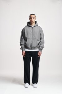 Young man standing facing forward wearing a grey zip-up hoodie, black pants, white sneakers, against a plain white background.