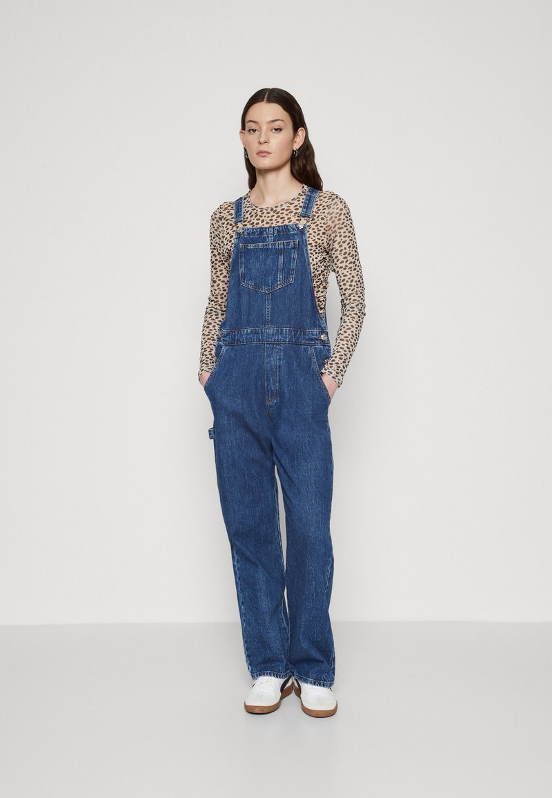 Denim naramnice temno modre barve z sproščenim krojem, ki vključujejo nastavljive naramnice, sprednji žep in stranske žepke, v kombinaciji z vzorčasto bluzo.