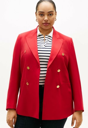 Vrouw die een rood dubbelbreasted blazer met gouden knopen draagt over een wit- en marineblauw gestreept shirt en donkere broek, tegen een witte achtergrond.