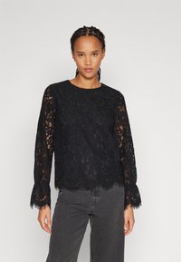 Black lace top with long sleeves, scalloped hem, and fringe accents. Lightweight, sheer fabric with floral patterns. Paired with gray jeans.
