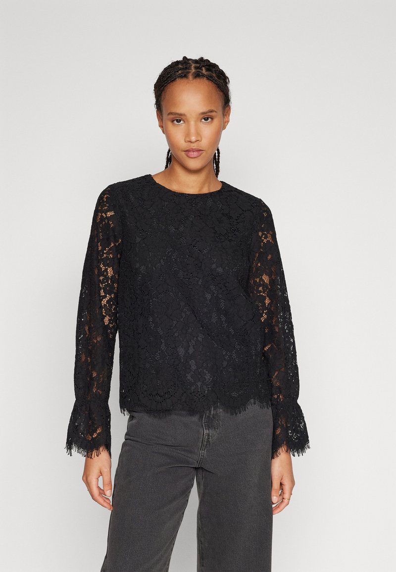 Black lace top with long sleeves, scalloped hem, and fringe accents. Lightweight, sheer fabric with floral patterns. Paired with gray jeans.