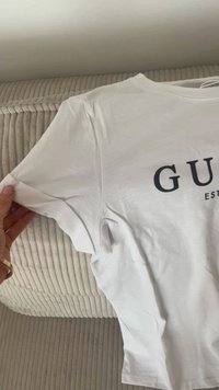 T-shirt bianco in cotone con maniche corte, caratterizzato da un logo nero con il testo "GU" e "EST" sottostante. Tessuto morbido con una vestibilità casual.