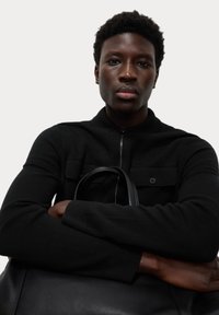 Sort zip-up sweater med knaplommer på brystet og ribbede cuffs; modellen holder en sort lædertaske med glat tekstur.