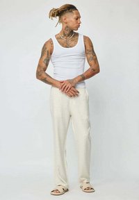 Débardeur blanc sans manches associé à un pantalon texturé de couleur crème et des sandales à glissière beige clair. Le mannequin présente de nombreux tatouages.