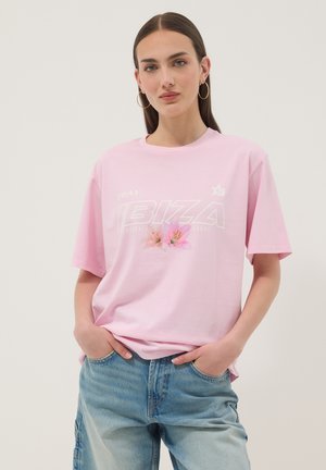 Joven mujer con largo cabello castaño lleva pendientes de aro dorados, una camiseta rosa claro "IBIZA" con diseño floral y vaqueros azules con las manos en los bolsillos.