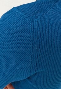 Blå stickad tröja med ett texturerat ribbat mönster och tydliga axelsömmar, som visar en närbild av tygets form.