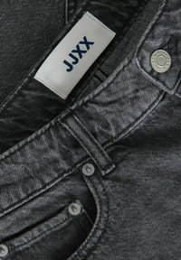 Tejido de mezclilla de color gris oscuro con una superficie ligeramente texturizada. Presenta una etiqueta blanca con "JYXX" y herrajes de botón metálico.