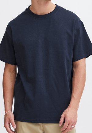 Mand iført ensfarvet mørkeblå T-shirt med korte ærmer og beige bukser, der viser overkroppen og arme uden synligt ansigt.