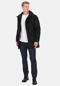 Giacca imbottita nera con cappuccio, dotata di tasche con cerniera e design elegante. Abbinata a una camicia bianca e jeans in denim scuro.