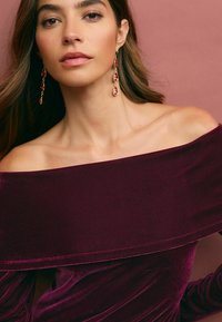 Burgundy sammet off-shoulder topp med en mjuk textur, draperad design och accentuerad med långa örhängen i guld och röd.