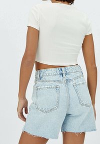 Mujer vestida con shorts de mezclilla azul claro y una camiseta corta de manga corta blanca de canalé, vista desde atrás contra un fondo liso.