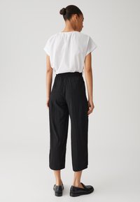 Zwarte cropped broeken met textuurstof en een elastische tailleband, gedragen met een wit T-shirt met korte mouwen. Er worden zwarte instapschoenen gedragen.