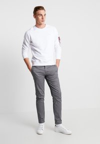 Felpa bianca in cotone con polsini a costine, abbinata a pantaloni slim fit grigi. Il modello indossa sneakers bianche. Design semplice e pulito.