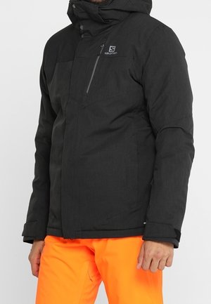 Homme portant une veste d'hiver noire Salomon avec capuche et un pantalon orange vif, debout devant un fond uni.