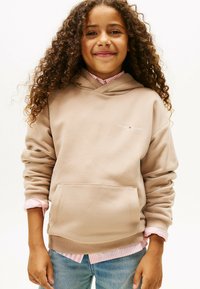 Niño sonriente con cabello rizado largo, lleva puesto un sudadera beige de Tommy Hilfiger sobre una camiseta de rayas rosas y jeans azules claros, de pie contra un fondo blanco.