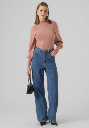 Maglione aderente rosa con maniche a sbuffo, abbinato a jeans blu a vita alta e a gamba larga. La modella tiene una piccola borsa nera. Sfondo grigio chiaro.