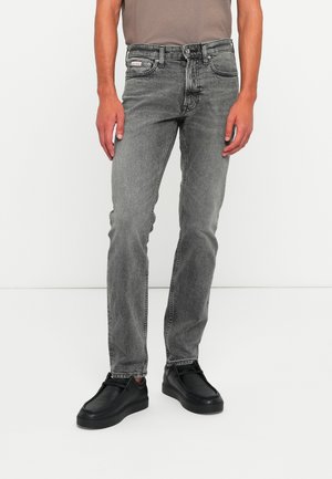 Man wearing gray slim-fit jeans and black leather shoes standing against a plain white background.