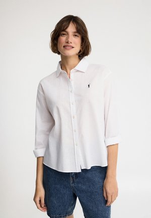 Kobieta o krótkich brązowych włosach nosi białą koszulę na guziki z podwiniętymi rękawami i ciemnoniebieskie szorty jeansowe, stoi na jednolitym tle.