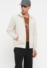 Άνδρας με γκρι καπέλο, κρεμ ζακέτα με φερμουάρ, καφέ μπλούζα Tommy Hilfiger και μαύρο παντελόνι, στέκεται με το ένα χέρι στην τσέπη.