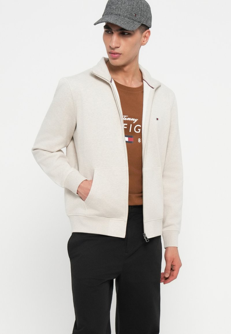 Mann trägt eine graue Kappe, eine cremefarbene Zip-Jacke, ein braunes Tommy Hilfiger-Shirt und schwarze Hosen, steht mit einer Hand in der Tasche.